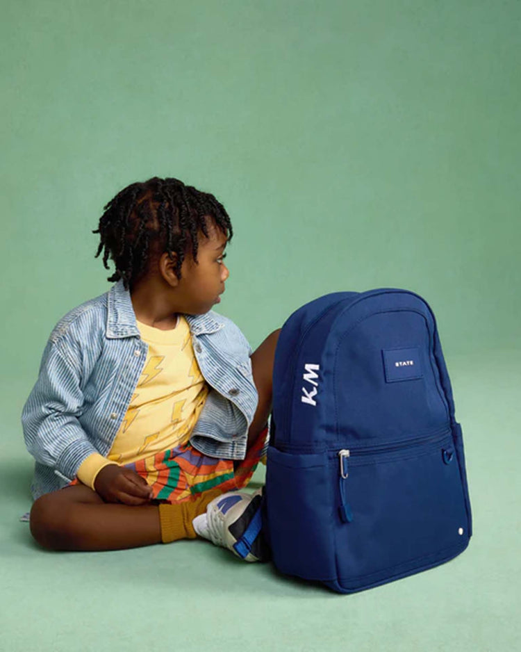 A young child sits cross-legged on the floor, looking to the side, with a large navy STATE Bags Kane Mini Backpack labeled KM in front of them. The background is a solid light green color.