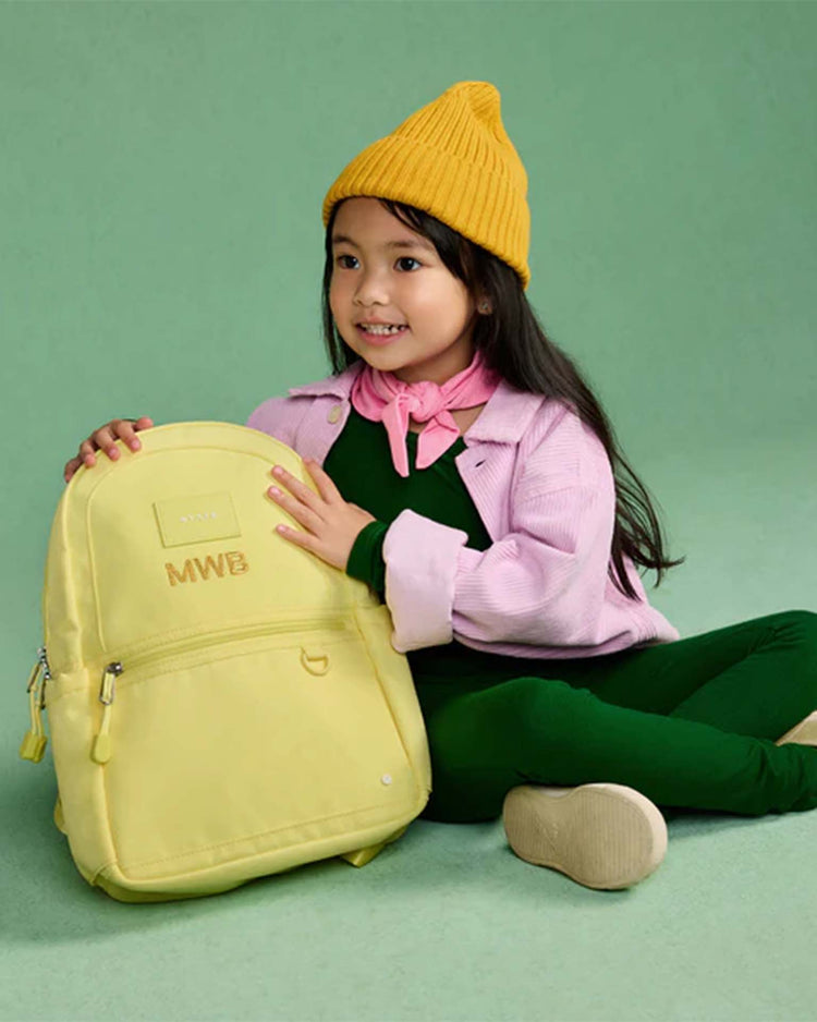 A young girl in a yellow beanie and pink jacket sits on the floor, smiling and holding a State Bags Kane Mini Backpack in Butter Yellow, against a green background.