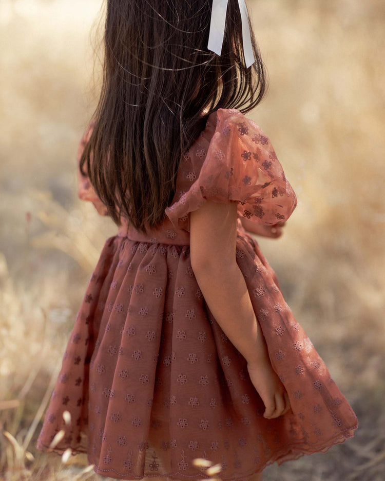 A young girl with long brown hair tied with a white ribbon stands outdoors in a sunlit, grassy field, wearing the Noralee “Sofia” dress in Spice Floral—a puff-sleeve pink organza dress adorned with delicate floral patterns. She faces away from the camera.