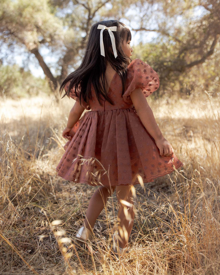 A young girl with long dark hair and a white bow wears the noralee sofia dress in spice floral as she stands in a dry, grassy field surrounded by trees, looking away from the camera.