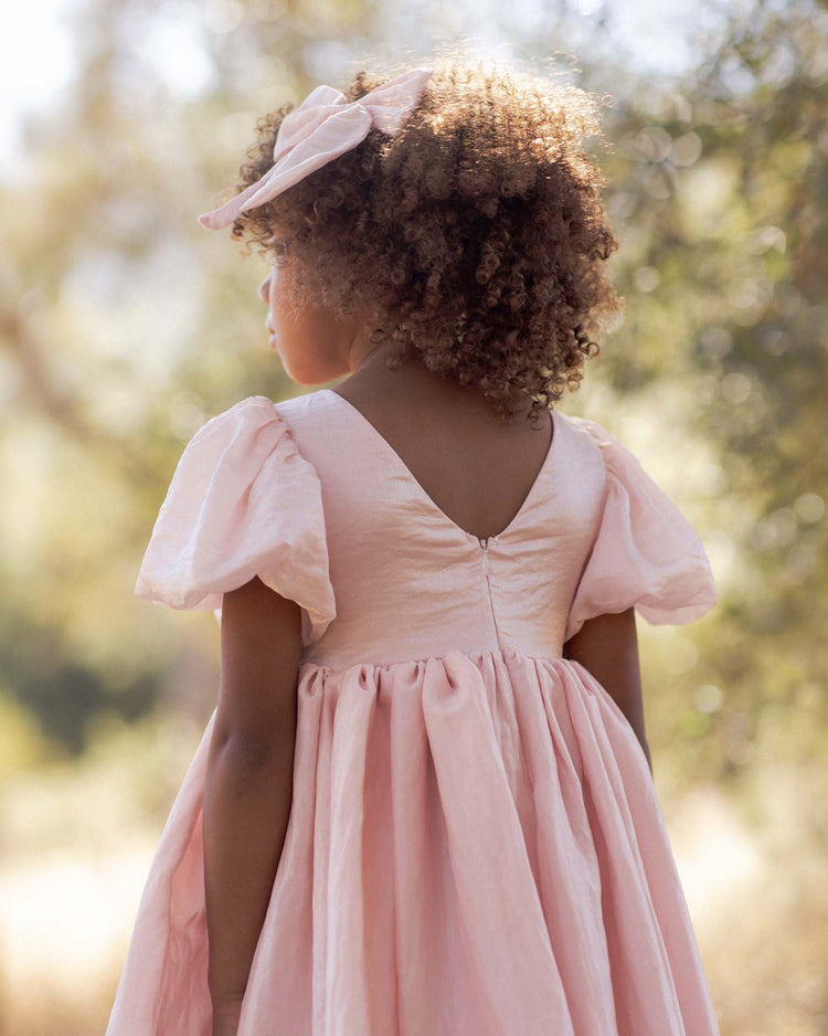 A young girl with curly hair stands outdoors, facing away, wearing the noralee sofia dress in rose shimmer with a matching headband; trees and sunlight fill the background.