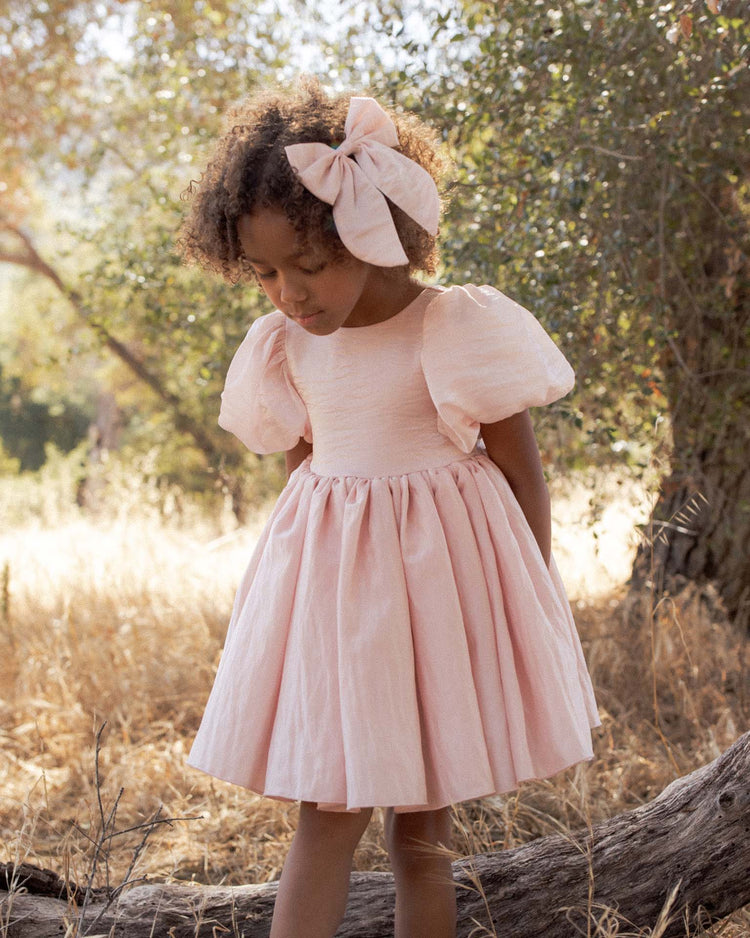 A young girl wearing the noralee sofia dress in rose shimmer and a matching large bow stands outdoors in a sunlit field with trees and dry grass in the background.