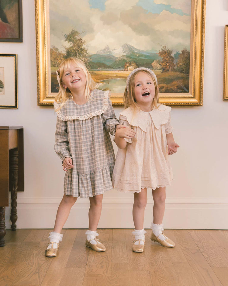 Two young girls stand smiling and holding hands in a room with wooden floors and artwork on the walls. One wears the “noelle dress in fall plaid” by noralee, featuring a sailor bib collar, while both pair their light-colored dresses with white socks and gold shoes.