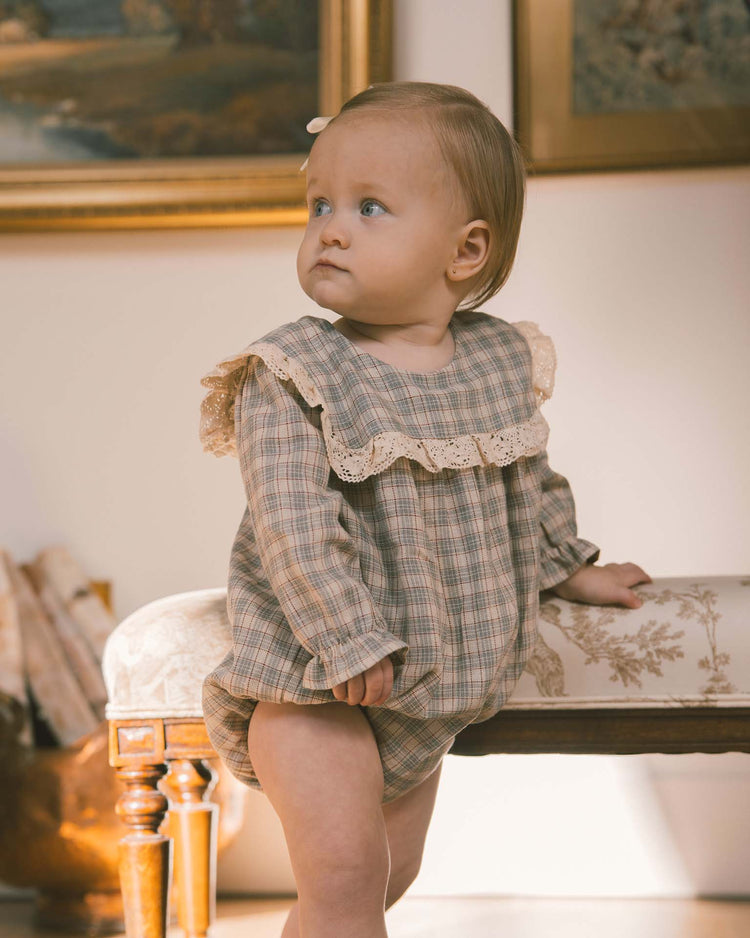A baby, dressed in the Noralee noelle romper in fall plaid with its charming lace collar, stands holding onto a cushioned bench. With a hair clip in place, the baby gazes to the side in a warmly lit room adorned with framed art.