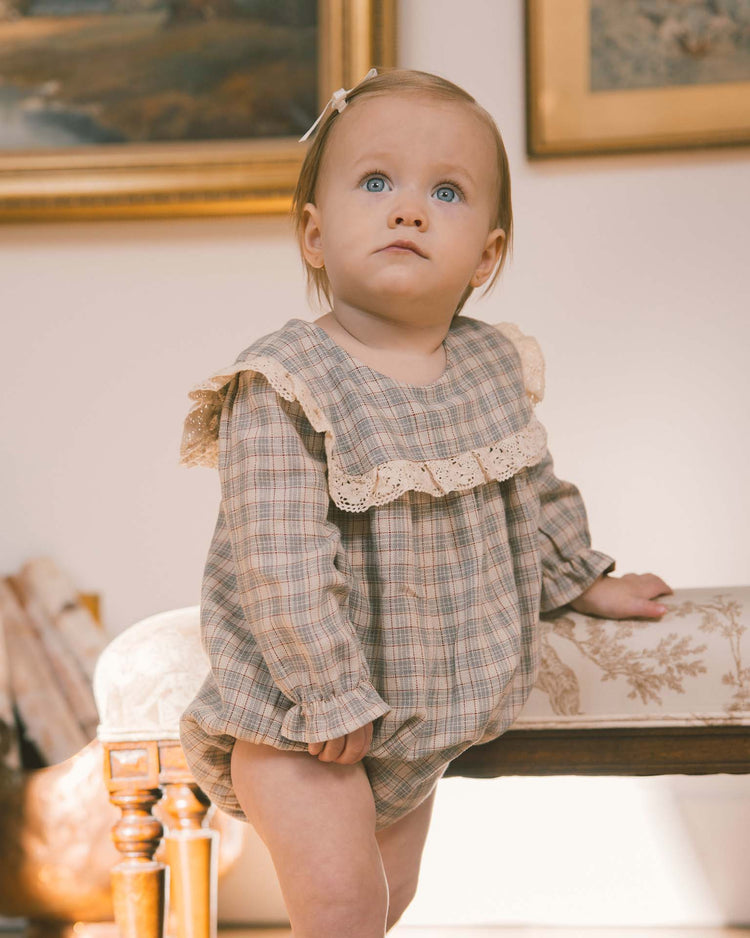 A baby with blue eyes and light brown hair wears the Noralee Noelle Romper in Fall Plaid, a charming piece featuring delicate lace trim, while sitting on a cushioned bench in a softly lit room adorned with framed artwork.