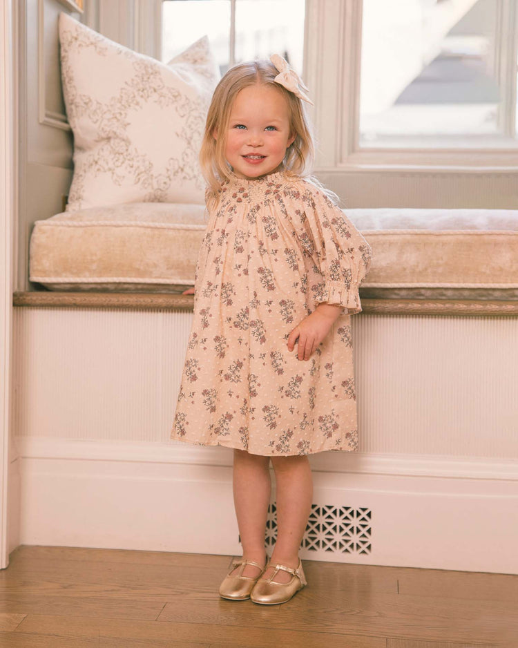 A young girl with blonde hair stands smiling in front of a window seat with cream cushions in a softly lit room, wearing noralee’s Maddie Dress in French Floral—crafted from delicate swiss dot fabric—and subtle gold shoes.