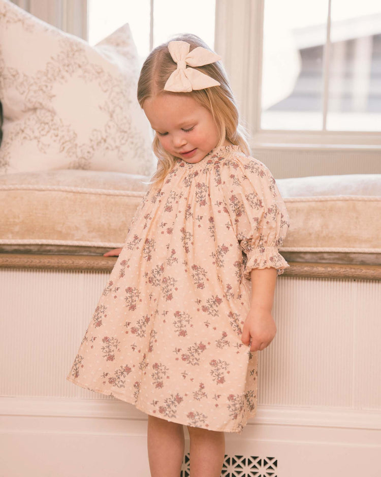 A young child with a cream bow in her hair wears the noralee “maddie dress in french floral,” featuring delicate light pink floral embroidery, while looking down and standing indoors near a window seat with a patterned cushion.