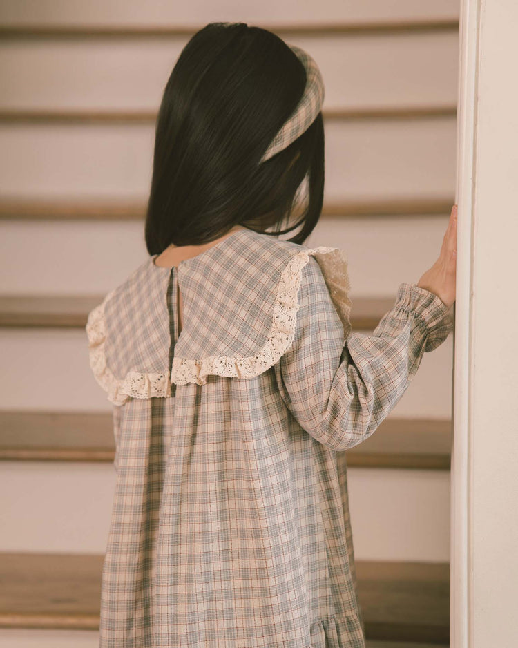 A young girl with long dark hair, wearing the noralee noelle dress in fall plaid with a sailor bib collar, lace trim, and a matching headband, stands on a staircase facing away while holding the railing.