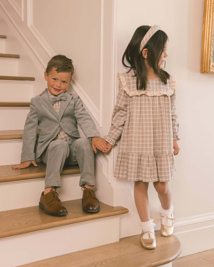 A boy in a light suit and brown shoes sits on stairs, holding hands with a girl beside him dressed in the noralee noelle dress in fall plaid with a coordinating headband. Both children appear happy and stylish.