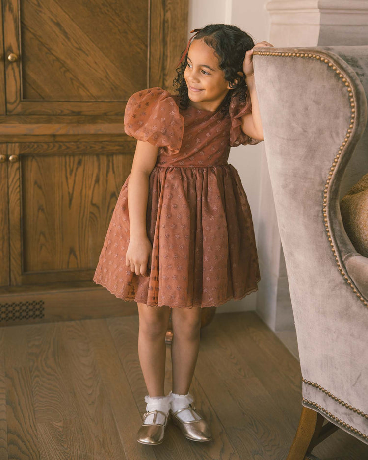A young girl in the noralee sofia dress in spice floral, featuring balloon puff sleeves, and gold shoes leans on a gray armchair with a gentle smile. She stands on wooden flooring beside a wooden cabinet.