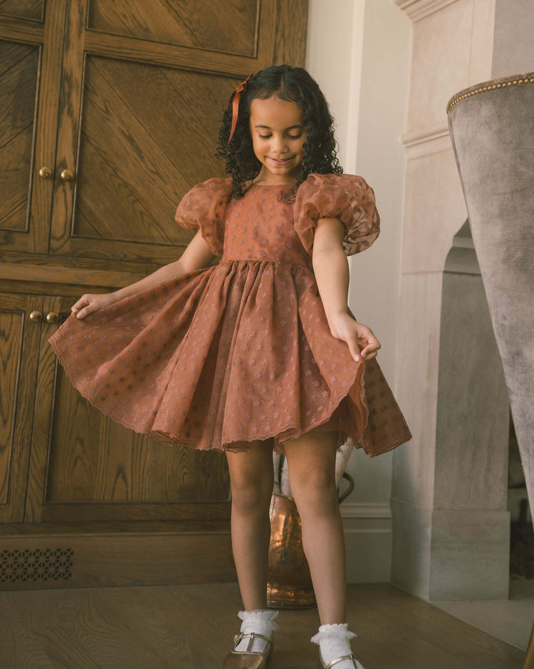 A young girl with curly hair wears the noralee sofia dress in spice floral, featuring balloon puff sleeves, paired with white shoes as she stands indoors holding out the sides of her dress and smiling down.