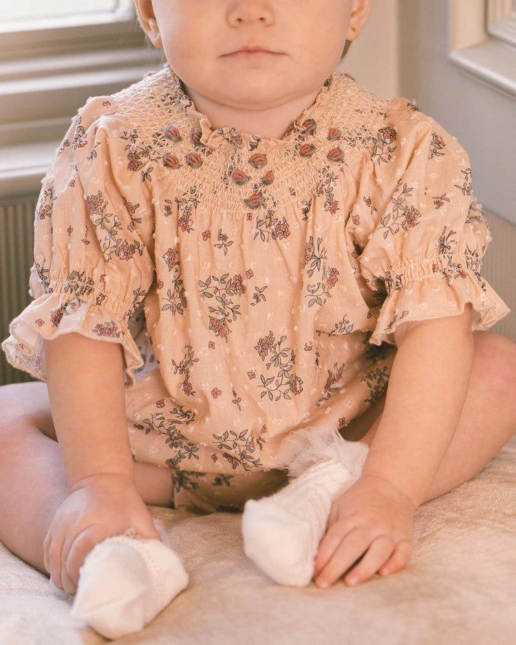 A baby wearing the noralee maddie romper in french floral, crafted from delicate swiss dot fabric with charming floral embroidery, sits on a beige surface near a window. The baby’s face is partially visible, and their hands rest gently on their legs, complemented by white socks.