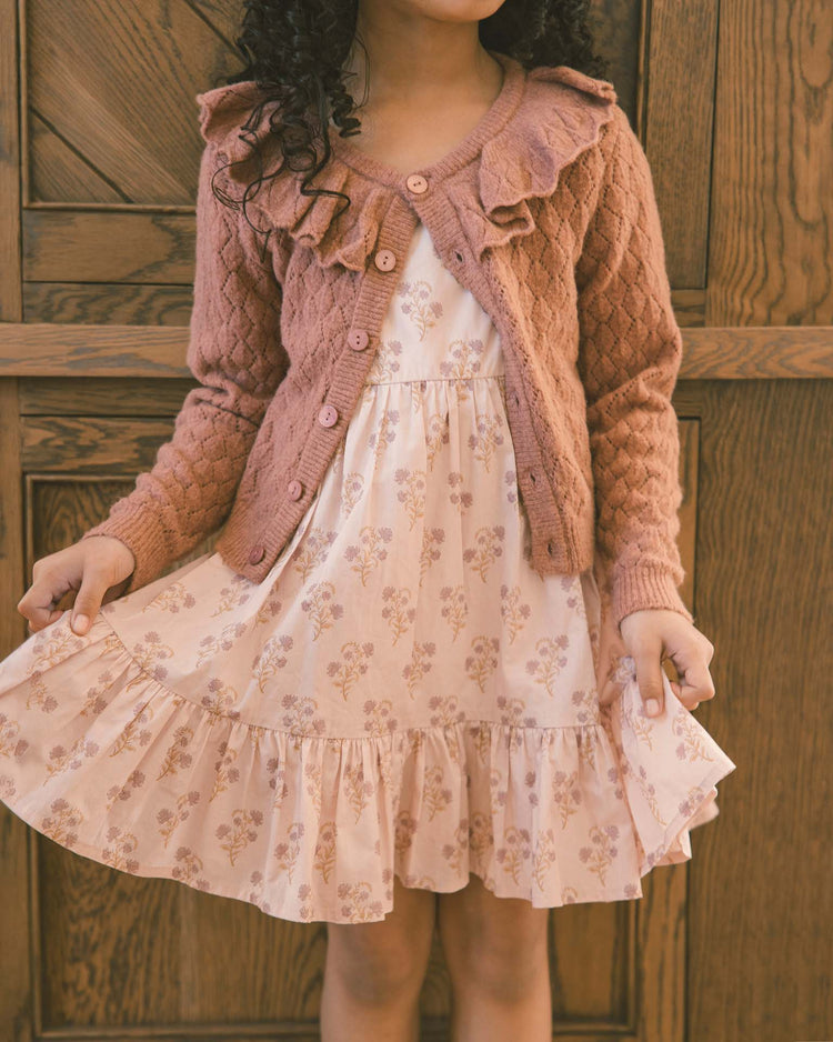 A young girl wearing the noralee ruffle cardigan sweater in spice with an oversized collar and a pink floral dress stands in front of a wooden door, holding out the sides of her dress.