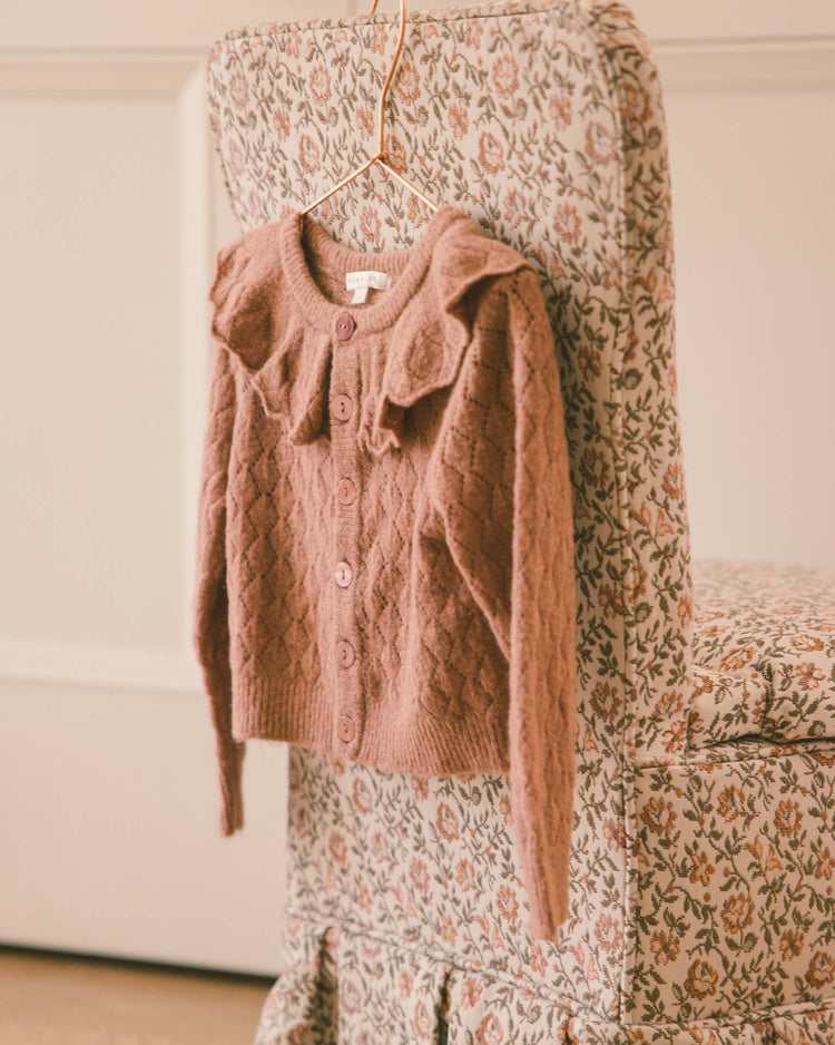 A spice-colored, pointelle knit children’s ruffle cardigan sweater from noralee hangs on a copper hanger, displayed on a floral-patterned chair with a matching fabric cover and ruffled skirt.