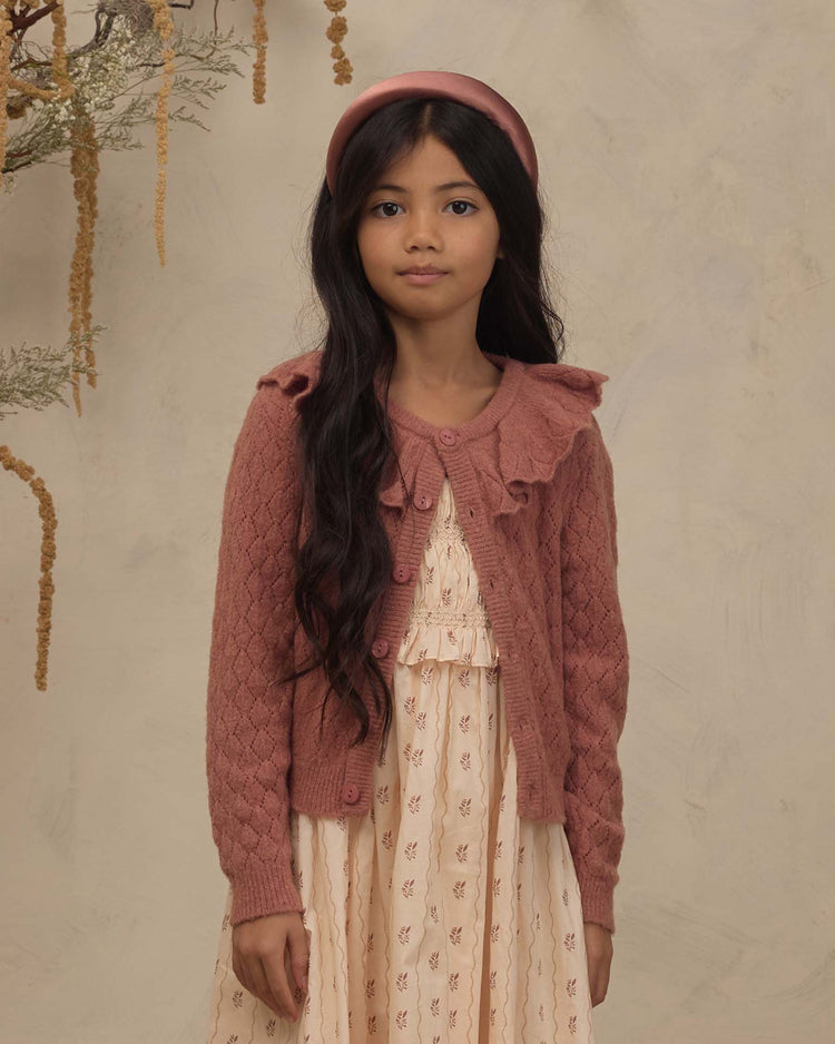 A young girl with long dark hair, wearing a pink headband and the noralee ruffle cardigan sweater in spice layered over a cream printed dress, stands indoors against a neutral backdrop adorned with hanging plants.