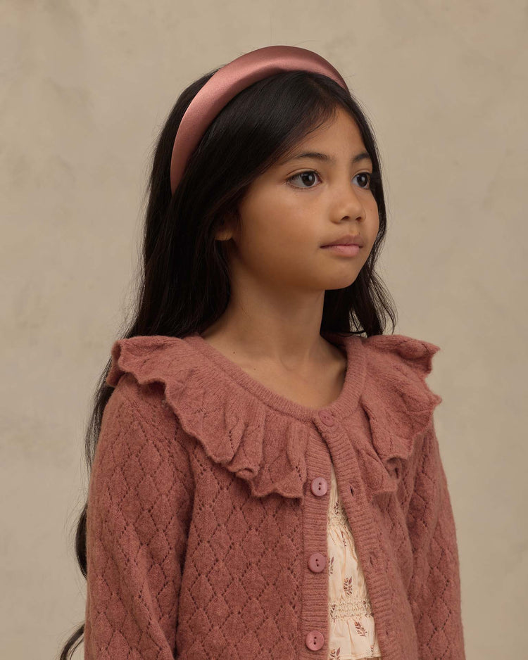 A young girl with long dark hair, wearing a pink headband and the noralee ruffle cardigan sweater in spice—a textured piece featuring a ruffled collar—stands against a neutral background, gazing slightly to the right.