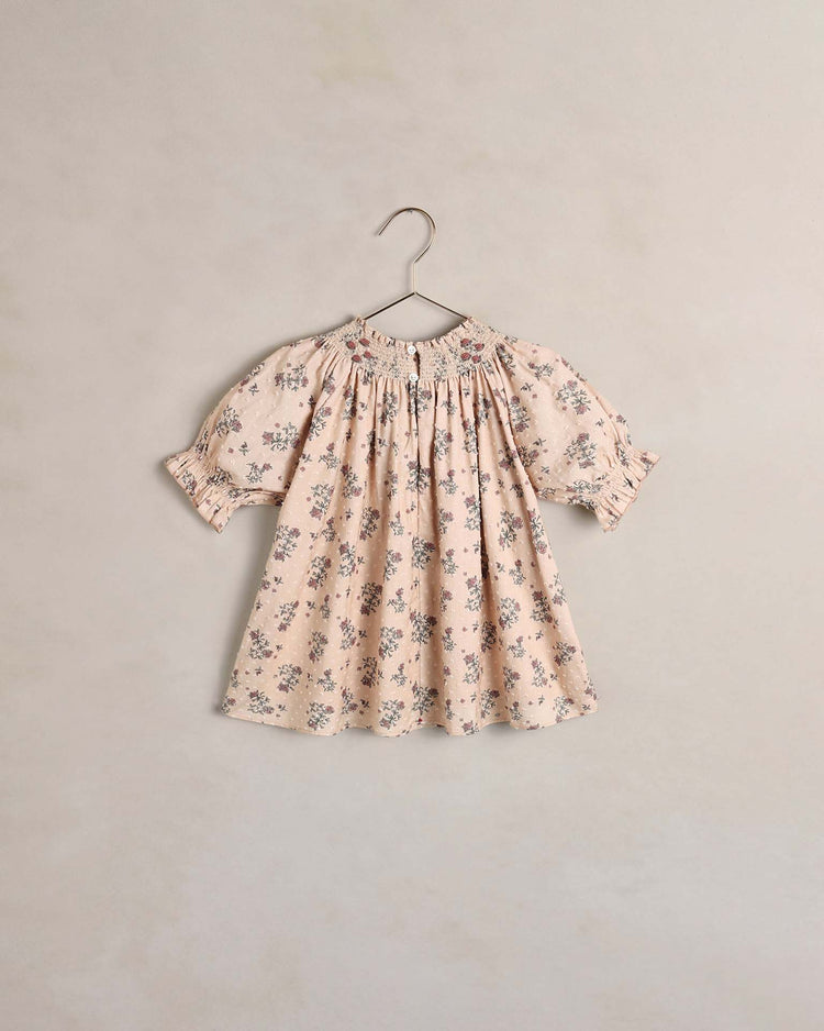 The noralee maddie dress in french floral, a short-sleeved baby dress in pale pink with a delicate floral pattern, is crafted from swiss dot fabric and hangs on a metal hanger against a plain, light-colored wall. This vintage-inspired piece infuses timeless charm into any little one’s wardrobe.