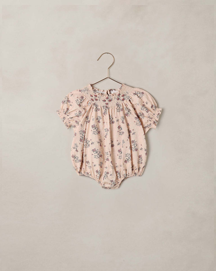 The “maddie romper in french floral” by noralee—a pink vintage-inspired romper with short sleeves, delicate floral print, elastic at the legs and sleeves, and charming floral embroidery—hangs on a gold hanger against a neutral background.