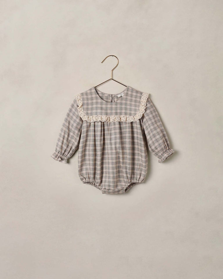 The noelle romper in fall plaid by noralee is a long-sleeved gray baby romper adorned with delicate lace trim across the chest and ruffled sleeve cuffs, elegantly showcased on a minimalist metal hanger against a plain beige backdrop.