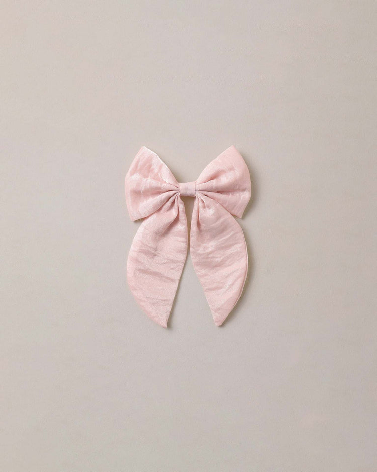 A large everly bow in rose shimmer by noralee, featuring two long tails and a soft pale pink hue with a subtle shimmer, is displayed against a plain light gray background.