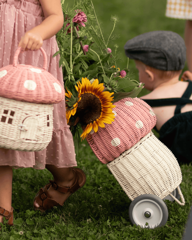 Two children outdoors: one in a pink dress pulls a dinkum dolls rattan mushroom luggy in musk, filled with flowers including a sunflower, and holds a matching mushroom-shaped basket. Another child sits on the grass behind her.