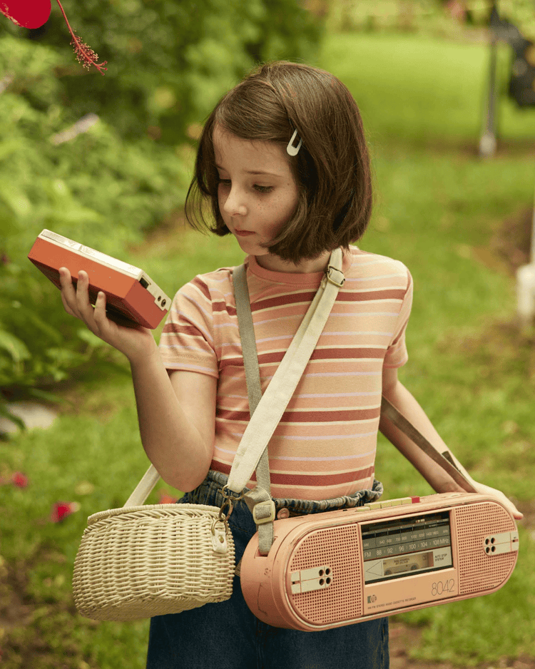 A young girl with a bob haircut stands outdoors in a striped shirt and jeans, holding a cassette tape and carrying the dinkum dolls mini chari bag in chalk, with greenery in the background.