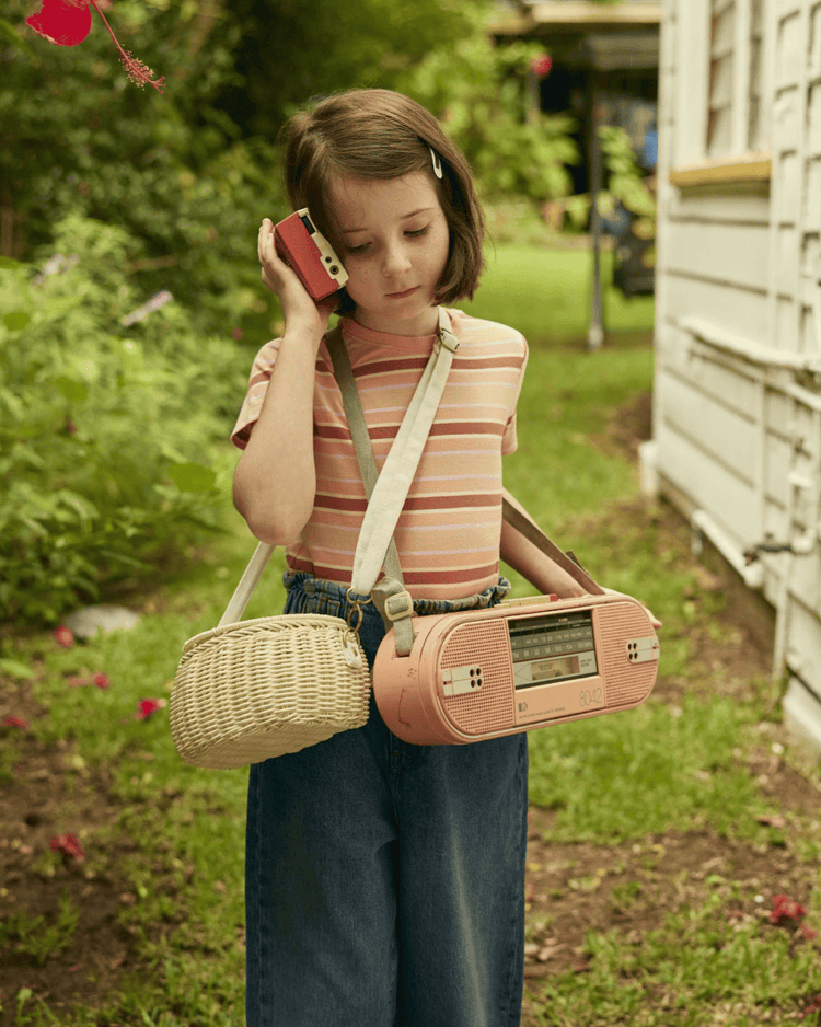 A young girl with a bob haircut stands outside by a white house, holding a red cassette player and carrying the dinkum dolls mini chari bag in chalk; she wears a striped shirt, jeans, and the green background is leafy.
