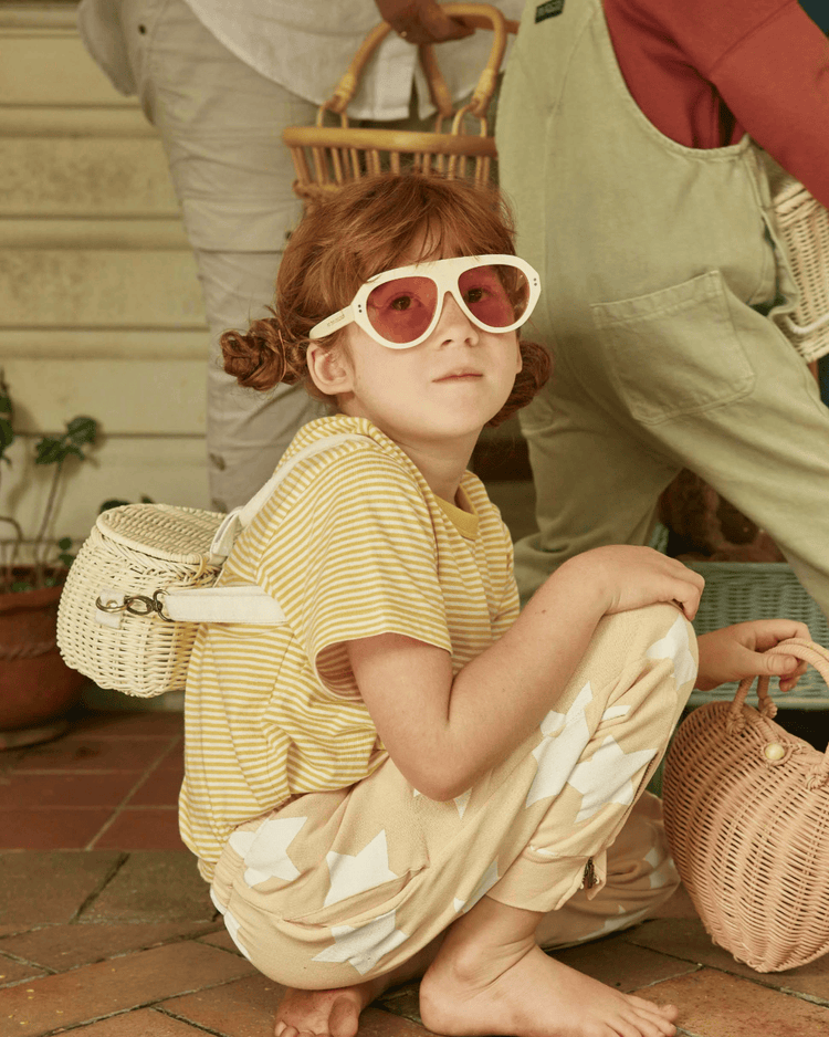 A young child with red hair crouches barefoot on a tiled porch, holding the dinkum dolls mini chari bag in chalk. Large round sunglasses, a yellow striped shirt, and patterned pants complete the look; adults and baskets appear in the background.