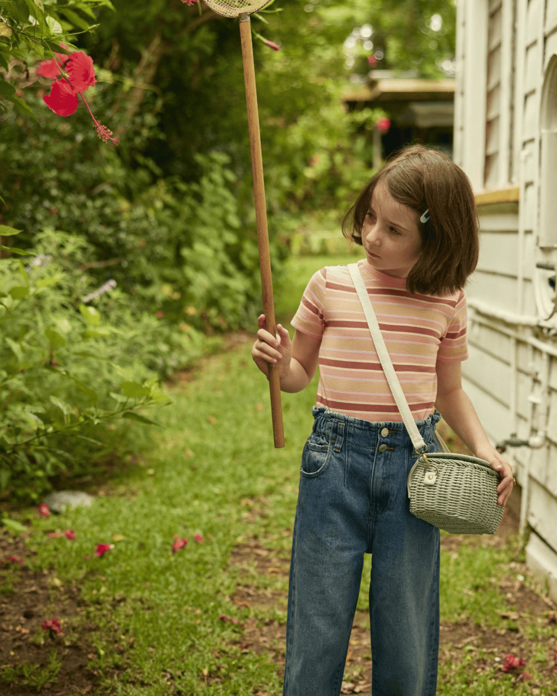A young girl with short brown hair stands in a garden, holding a net toward a pink hibiscus flower. She wears a striped shirt, blue jeans, and carries the dinkum dolls mini chari bag in vintage blue over her shoulder; a white house is on the right.