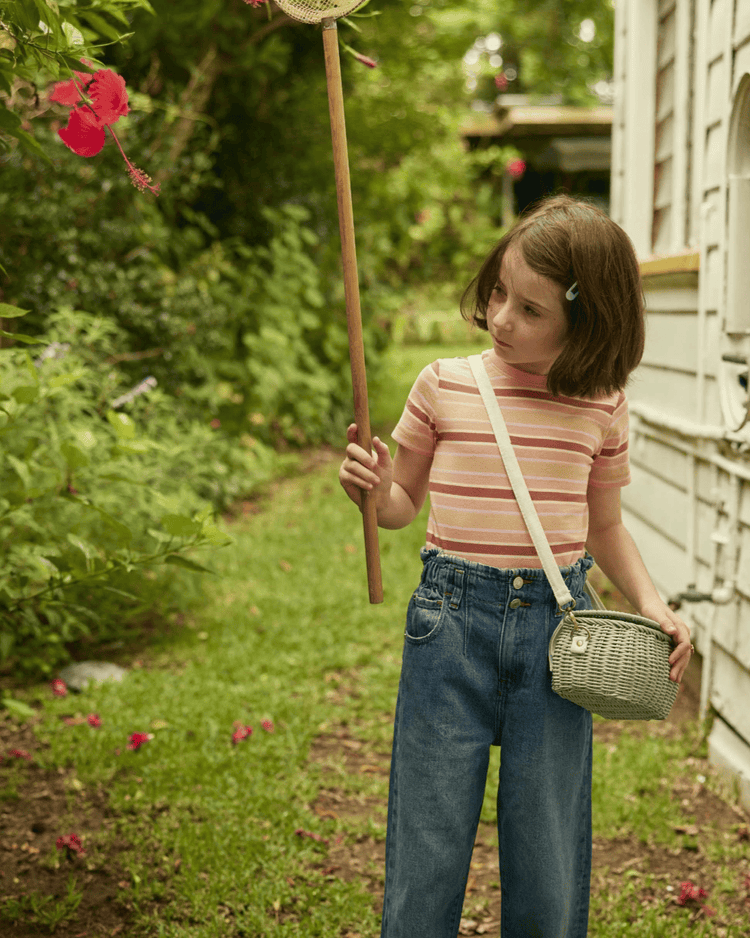A young girl with short brown hair stands in a garden, holding a net toward a pink hibiscus flower. She wears a striped shirt, blue jeans, and carries the dinkum dolls mini chari bag in vintage blue over her shoulder; a white house is on the right.