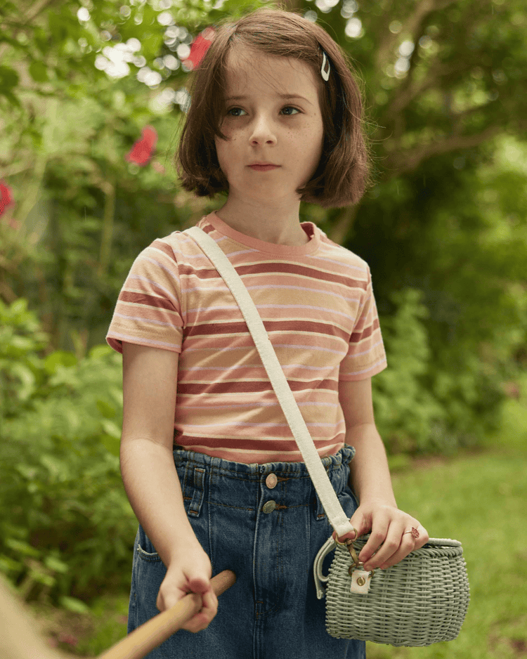 A young girl with short brown hair stands outdoors among greenery, holding a stick. She wears a pink and white striped t-shirt, high-waisted jeans, and carries a dinkum dolls mini chari bag in vintage blue over her shoulder.
