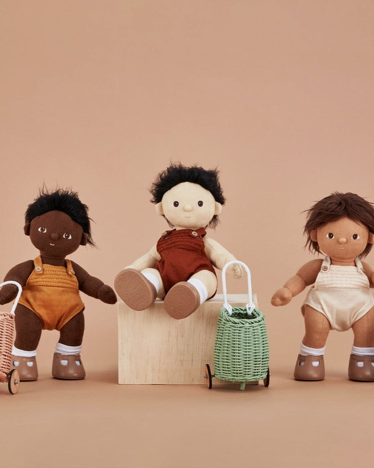 Three Dinkum Dolls with dark, medium, and light skin tones wear overalls and brown shoes. Two hold mint Doll Luggy baskets with wheels on a beige surface against a light brown background.