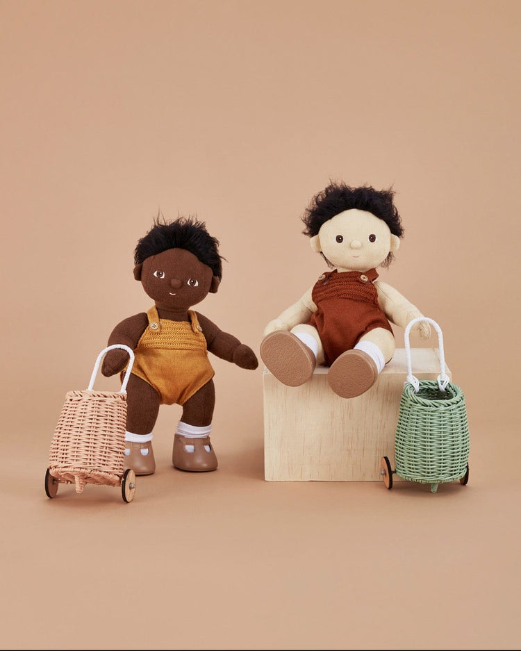 Two fabric Dinkum Dolls—one with dark brown skin in yellow overalls, one with light skin in rust—sit on a wooden block, each with a handwoven Doll Luggy in mint from dinkum dolls, against a beige background.