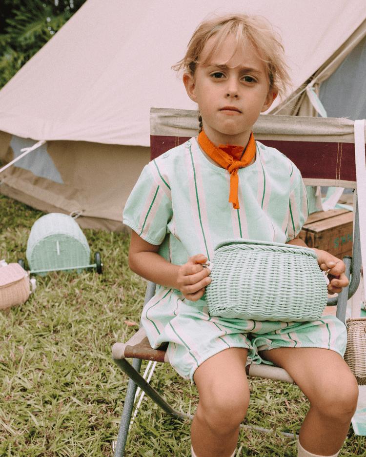 A young child with short light hair sits outdoors on a folding chair, wearing a light green striped outfit and orange neckerchief, holding a dinkum dolls mini chari bag in mint, with grass and a tent in the background.