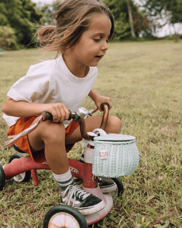 A young child with long hair rides a red tricycle on grass, wearing a white t-shirt, orange shorts, and sneakers. A dinkum dolls mini chari bag in mint is attached to the handlebars. Trees and greenery are in the background.