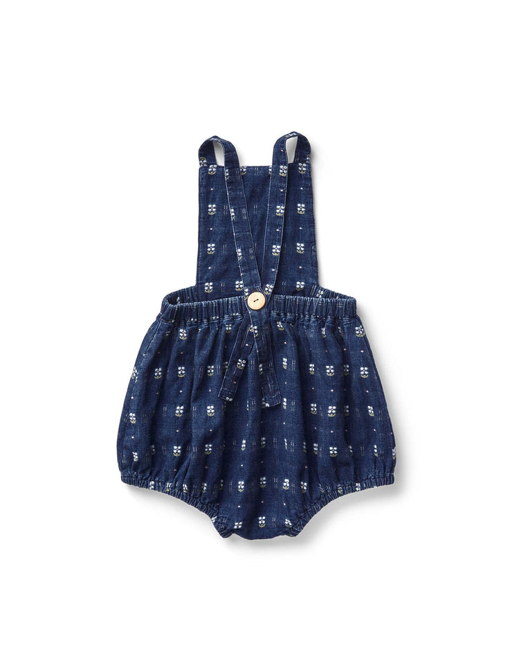 The oona romper in embroidered denim by soor ploom features an adorable white flower pattern, adjustable straps, and a front button. Made from soft cotton with care in Portugal, this charming piece is beautifully showcased on a white background.