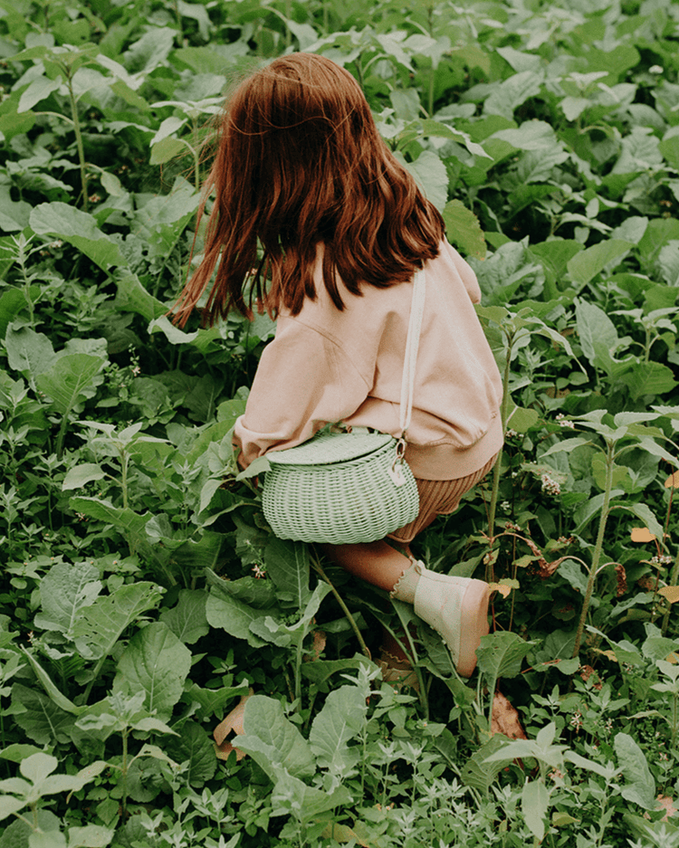 A young girl with brown hair, in a light sweater and skirt, kneels in a lush green field carrying the dinkum dolls mini chari bag in mint—a handwoven rattan purse—over her shoulder.
