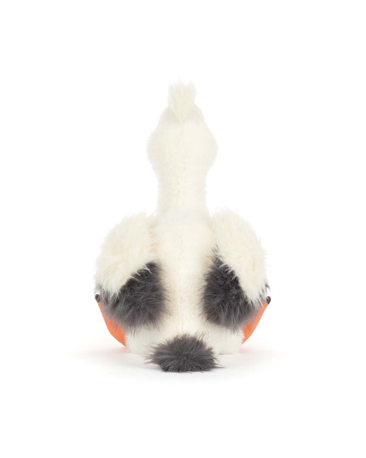 A Jellycat keli pelican plush toy sits with its back to the camera. It features white, black, and gray fur with orange accents on the sides and a fluffy gray tail, set against a plain white background.