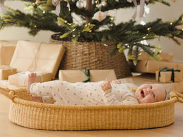 Baby lying in a woven basket in front of a decorated Christmas tree with presents underneath.