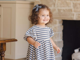 Child wearing a striped dress standing in a home setting with a fireplace.
