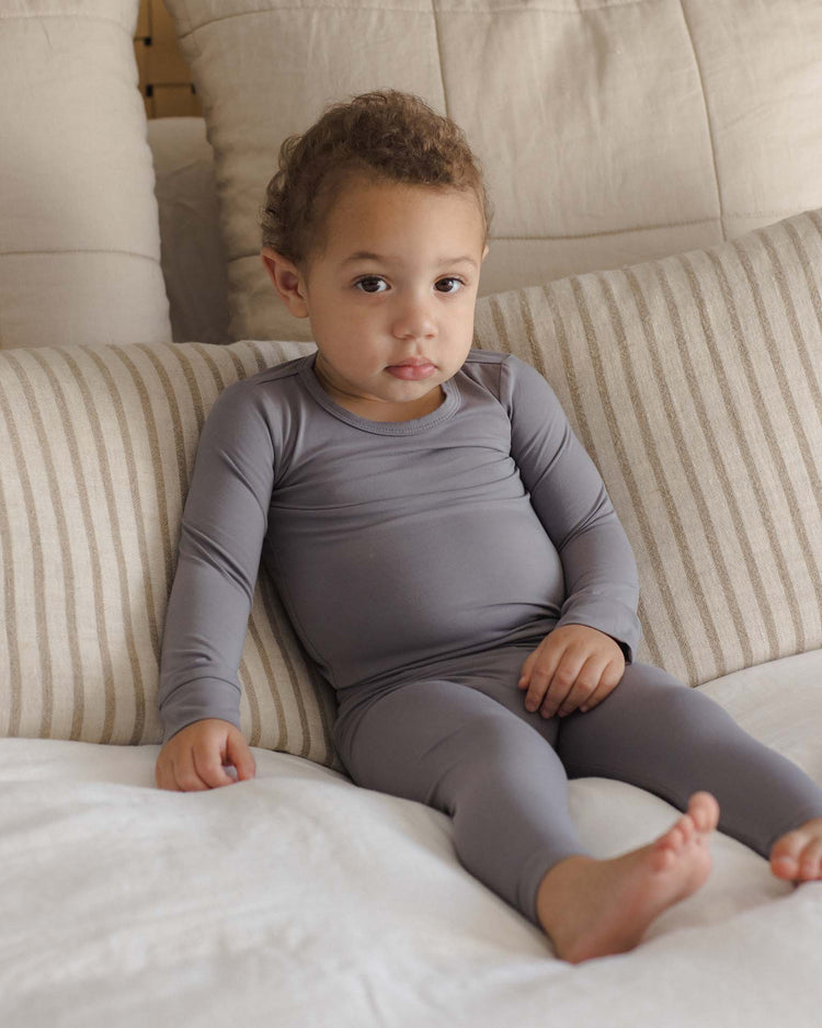 A young child with curly hair, wearing the quincy mae bamboo pajama set in slate, sits on a couch with beige striped pillows, looking directly at the camera.