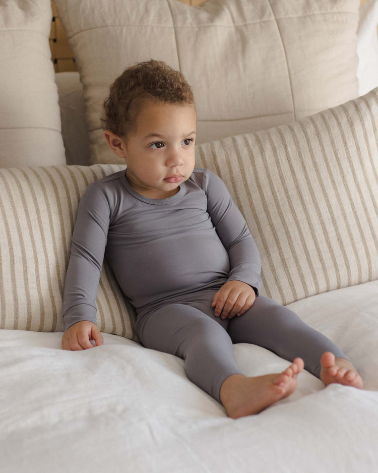 A young child wearing the quincy mae bamboo pajama set in slate sits on a bed with striped and solid pillows, gazing calmly to the side.