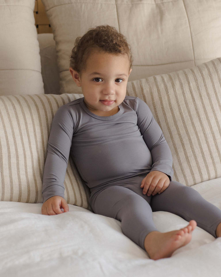 A young child with curly hair, wearing the quincy mae bamboo pajama set in slate, sits on a bed with striped and solid pillows, looking at the camera.
