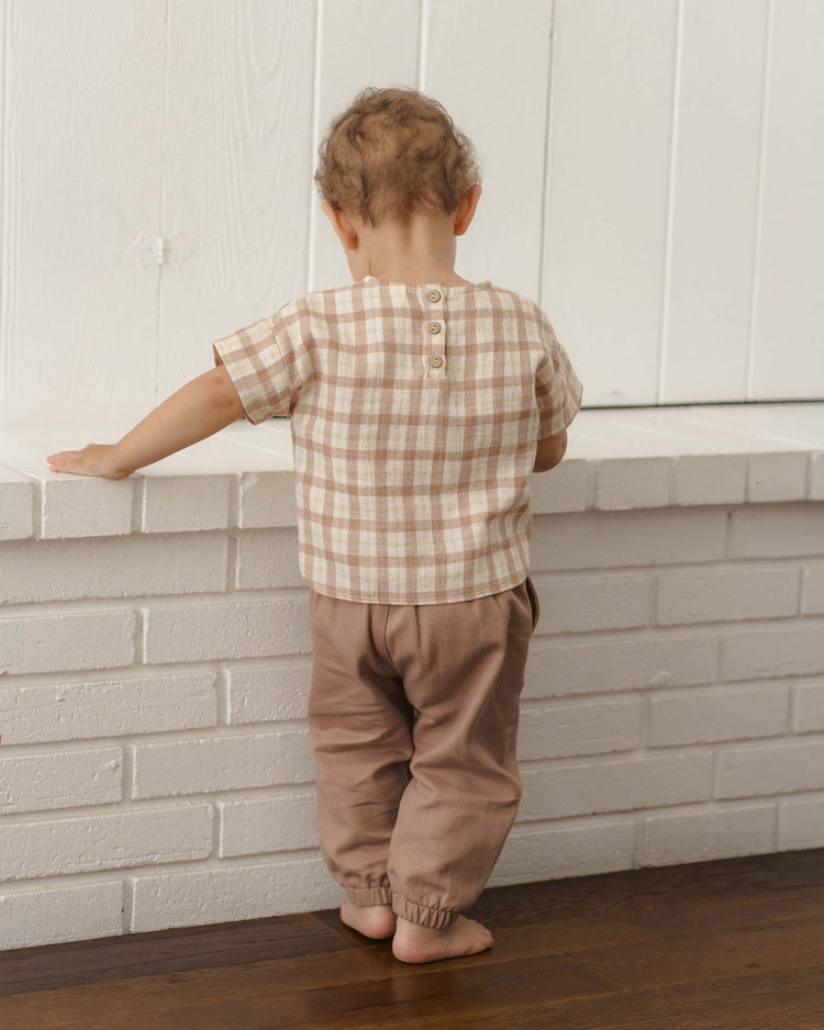 A toddler with curly hair, wearing the quincy mae woven boxy top in rustic plaid and light brown pants, stands facing a white brick wall with their back to the camera.