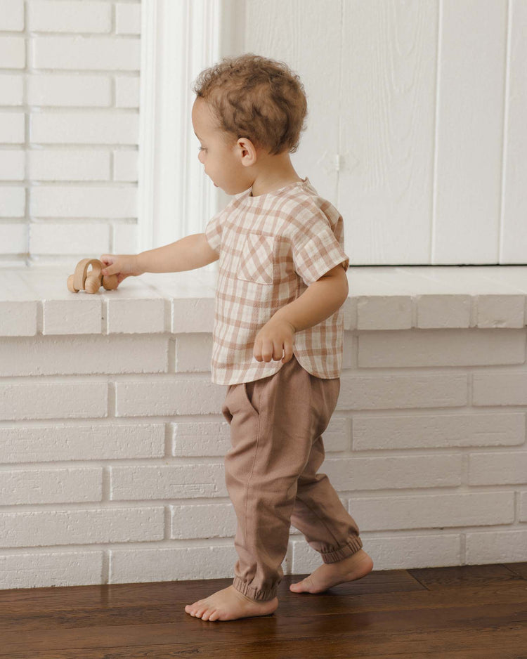 A toddler in a plaid shirt and quincy mae giovani pant in almond, featuring an elastic waistband and crafted from organic cotton, stands on a wooden floor, playing with a wooden toy on a white brick ledge.