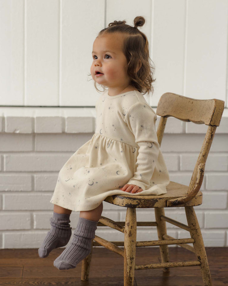 A toddler with brown hair in a topknot sits on a worn wooden chair, wearing a cream-colored dress with tiny patterns and cozy quincy mae socks from the socks set in slate + cloud. The background features a white brick wall and white wood paneling.