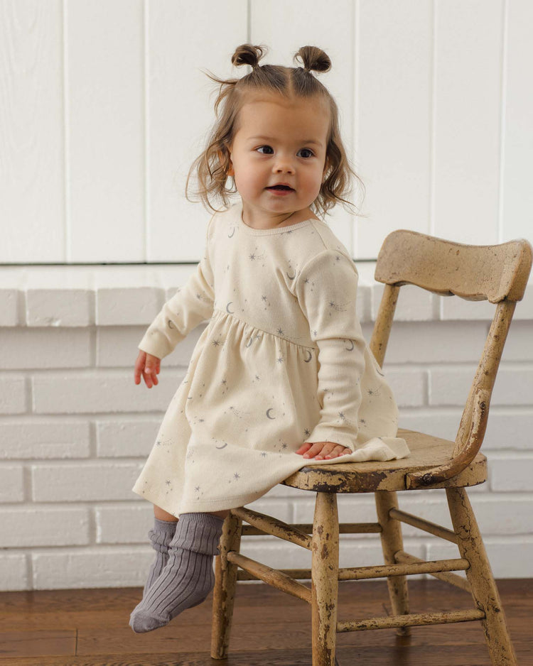 A toddler with brown hair in two buns sits on a rustic wooden chair, wearing a cream-colored dress with subtle patterns and cozy quincy mae socks from the socks set in slate + cloud, in front of a white brick wall.