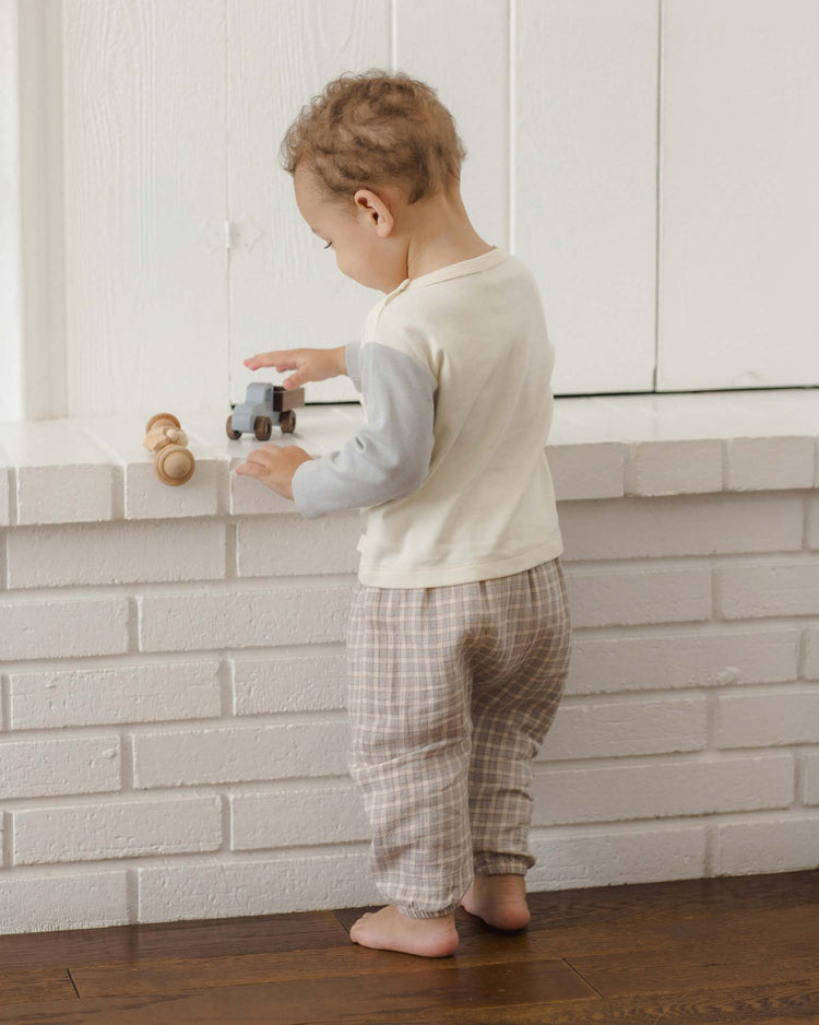 A toddler stands barefoot on a wooden floor, facing a white brick ledge, playing with two small wooden toy cars. The child wears plaid pants and the quincy mae long sleeve raglan tee in cloud, crafted from soft organic cotton.