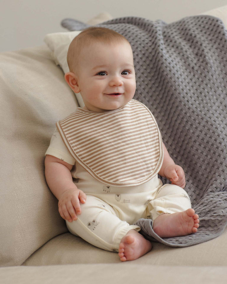 A smiling baby with light hair sits on a beige couch, wearing a cream outfit with animal prints and the quincy mae jersey snap bibs bunnies + almond stripe. A textured gray blanket is draped behind the baby.