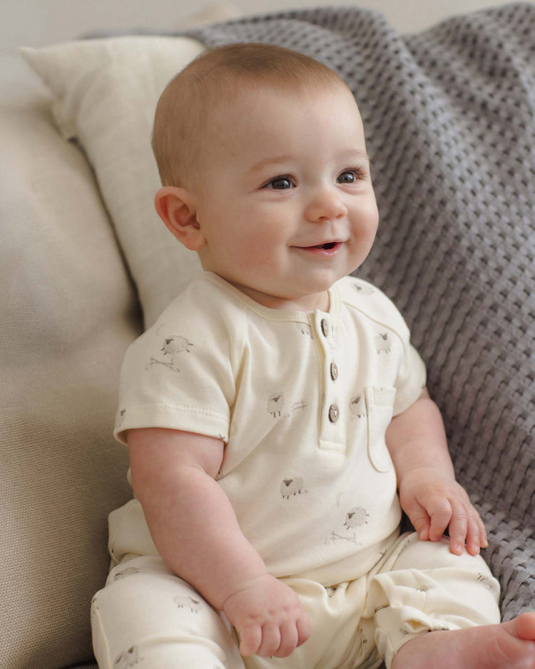 A smiling baby sits on a couch with beige and gray textured pillows, wearing the quincy mae short sleeve jumpsuit in sheep.